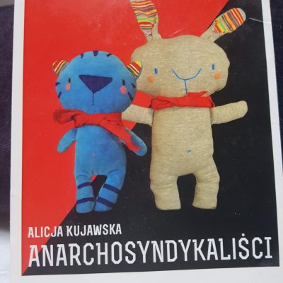 Alicja Kujawska, Anarchosyndykaliści, Zdjęcie Kartki Pocztowej 10,5x15,7 Cm., 2021 Zdjęcie Karty Pocztowej Towarzyszącej Wystawie Anarchosyndykaliści Z 2013 Roku. Na Pocztówce Znajduje Się Tytuł Anarchosyndykaliści I Nazwisko Autorki „Alicja Kujawska” Białymi Literami, Z Wielkich Liter. Tekst „Galeria 011 Od Nowa, 10.04.2013, Wernisaż 19" Zapisany Jest Mniejszymi Czarnymi Literami Na Białym Tle. Obok, W Prawym Dolnym Rogu, Również Na Białym Tle, Widać Logo Galerii 011, Klubu Od Nowa Oraz Uniwersytetu Mikołaja Kopernika W Toruniu. Centralne Miejsce Na Zdjęciu Zajmuje Flaga Anarchosyndykalistyczna, Czerwono-czarna, Diagonalnie Przecinająca Obraz. Na Jej Tle Widzimy Postaci Tytułowych Anarchosyndykalistów: Tygrysa I Królika. Obie Postaci To Rzeźby Miękkie, Zabawki. Tygrys (oryginalna Zabawka Wielkości 18 Cm) Z Niebieskiego Aksamitu, Stylizowany Na Chibi Albo Piernikowego Ludka (małe I Pulchne Karykatury Postaci Ludzkiej), Ma Nieproporcjonalnie Do Płaskiej „twarzy” Duży Granatowy Filcowy Nos, Małe Haftowane Oczy. Paski Tygrysa Są Granatowe. Uszy Symetryczne, Zdobione Materiałem W Paski. Po Prawej Stronie Królik Z Jasznoszarego Materiału, Uszy Z Materiału W Paski, Krzywo Przyszyte, Pyszczek, Oczy I Pępek Wyszyte Niebieską Nicią (wielkość Oryginalnej Zabawki 23 Cm). Obie Postaci Mają Na Szyjach Czerwone Chustki-bandany, W Nawiązaniu Do Spółdzielczych Działań Anarchosyndykalistów W Hiszpanii I Dawnej Jugosławii. Wystawa (składająca Się Z Rzeźb Miękkich W Różnych Aranżacjach Na Tle Rysunków Przedstawiających Miasto Budowane Przez Spółdzielców) Stanowiła Część Cyklu Wystaw Anarchiści, Którym Kuratorowała Katarzyna Lewandowska.