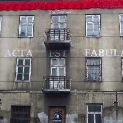 Anka Leśniak, Fifi Zastrow. Acta Est Fabula, Instalacja W Przestrzeni Publicznej, Łódź, 2015. Fotografia Instalację W Przestrzeni Publicznej Wykonaną Na Fasadzie Opuszczonego Budynku. Fasada Wypełnia Cały Kadr Zdjęcia. Pod Dachem Widać Czerwoną Kotarę Rozciągniętą Niemal Na Całej Szerokości Fasady. Pomiędzy Pierwszym A Drugim Piętrem, W Centralnej Części Fasady Znajduje Się Napis O Treści: Acta Est Fabula. Wykonany Został Krojem Pisma Antykwa. W środkowej Części Fasady Na Pierwszym I Drugim Piętrze Znajdują Się Drzwi Balkonowe Wraz Z Balkonami. Nad Drzwiami Balkonowymi Drugiego Piętra Widac Napis O Treści: Fifi Zastrow. Napis Wykonany Jest Ozdobnym Pismem Gotyckim Typu Szwabacha. Na Frontowych Drzwiach Zasłoniętych Drewnianymi Brązowymi żaluzjami Wisi Czarno-biały Plakat.