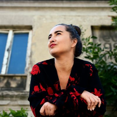 Anka Leśniak, zdjęcie portretowe, Fot. Agnieszka Cytacka, w ramach projektu Łodzianki. Atlas Piękna. Zdjęcie przedstawia kobietę - portret artystki patrzącej lekko w górę. Postać sfotografowana jest od dołu. Włosy ma splecione w kok. Ubrana jest w czarną bluzkę z czerwonymi kwiatami. W tle znajduje się fragment kamienicy z nieco zniszczoną fasadą oraz zielony krzew po prawej stronie kadru.