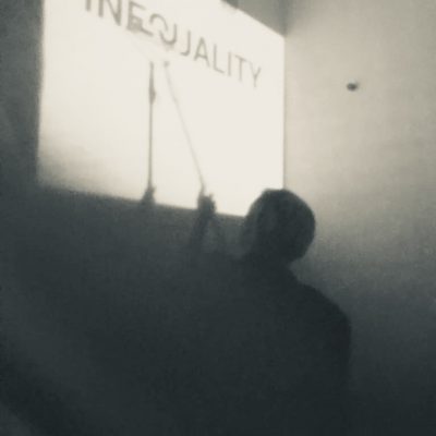 INEQUALITY Kadr Z Performance INEQUALITY. Czarno-białe Niewyraźne Zdjęcie Postaci, Artystki, Która Zamalowuje Wałkiem Z Białą Farbą, Białą ścianę, Na Której Jest Wyświetlony Jest Napis INEQUALITY – Nierówność.