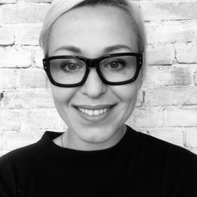 portret Moniki Czarskiej. Czarno-biały. Kobieta z blond włosami ubrana w czarną bluzkę stoi na tle ściany z cegieł. Uśmiecha się szeroko. Ma okulary z czarną oprawką.